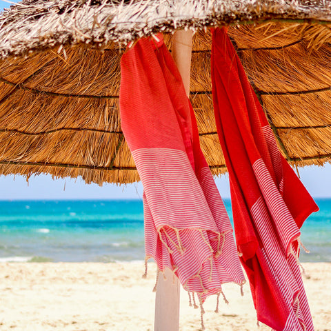 eine Korallen fouta und ein rotes Strandtuch unter einem Sonnenschirm am Strand