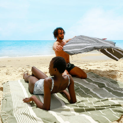 eine Frau am Strand, die auf einem olivgrünen Strandtuch liegt, und ein Mann der eine graue Fouta hält