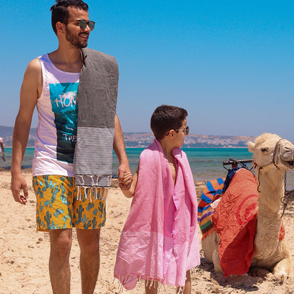 ein Vater in dunkelgrauer Fouta und der Sohn in rosa Fouta in der Nähe des Kamels am Strand